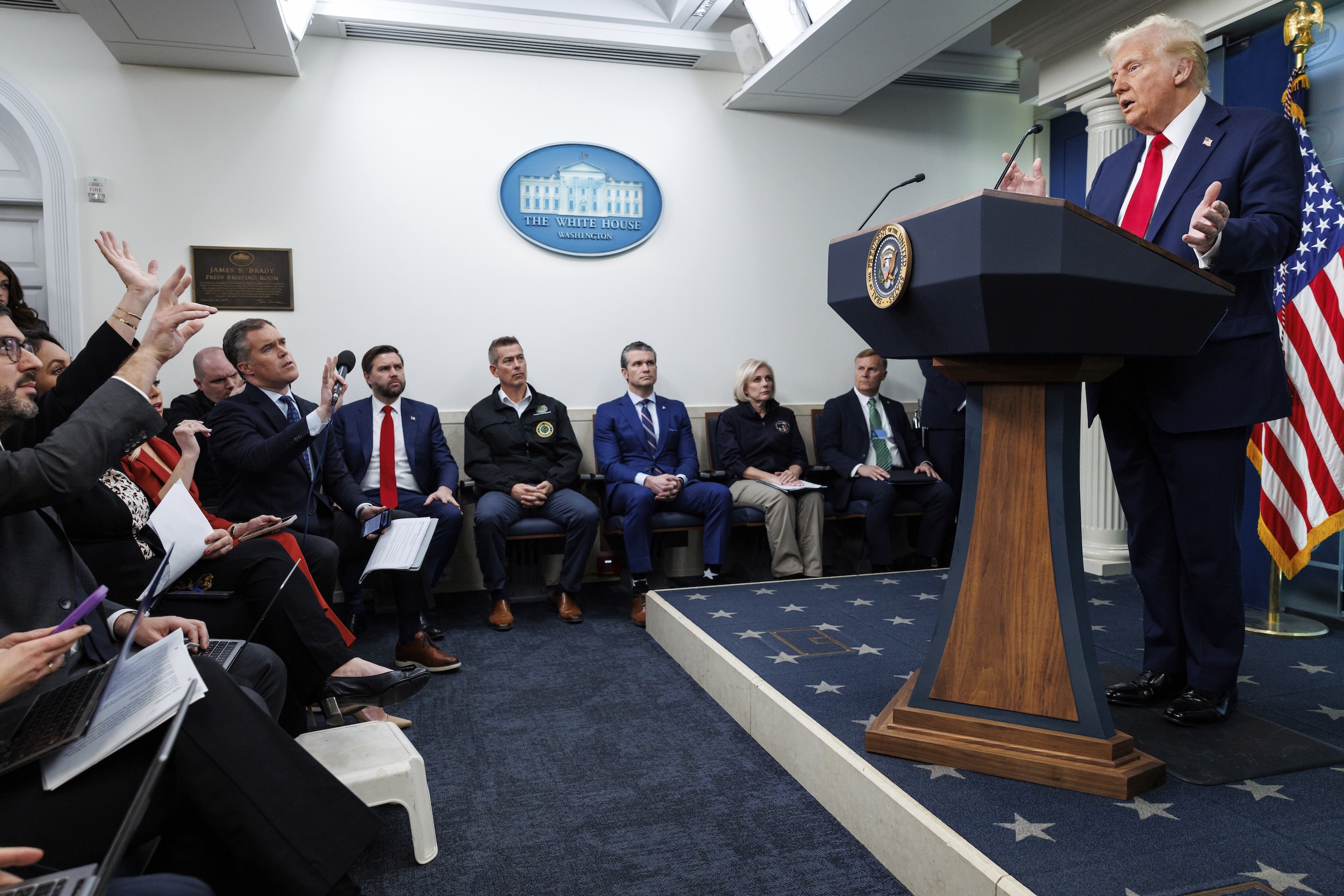 This image depicts President Donald Trump addressing a press briefing at the White House. He is standing at a podium with the presidential seal, speaking to an audience of journalists seated in the foreground and officials seated in the background. The journalists appear engaged, raising their hands and holding microphones or notebooks. The White House emblem and U.S. flag are visible in the room.