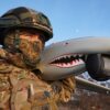 A Ukrainian soldier holds a Ukrspecsystems Shark UAV over his shoulder against a blue sky